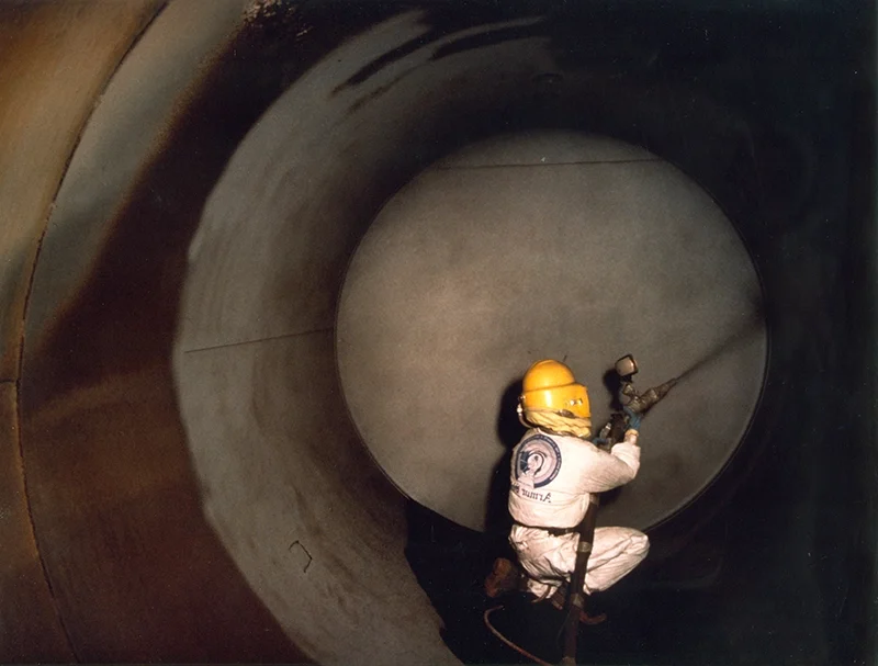 Crew member in protective gear sandblasting tank interior.